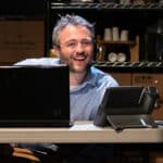 Alameda Post - An actor in Fully Committed sits behind a desk wearing a call headset and smiles.