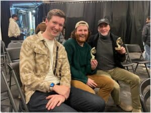Alameda Post - A photo of three men smiling and sitting together. Two hold small trophies.