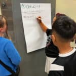 Alameda Post - Three fourth graders stand around a large piece of paper attached to the wall and work on a math problem together.