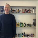 Alameda Post - A person in a blue sweater with short blond hair smiles at the camera in front of a wall of tattoo ink colors.