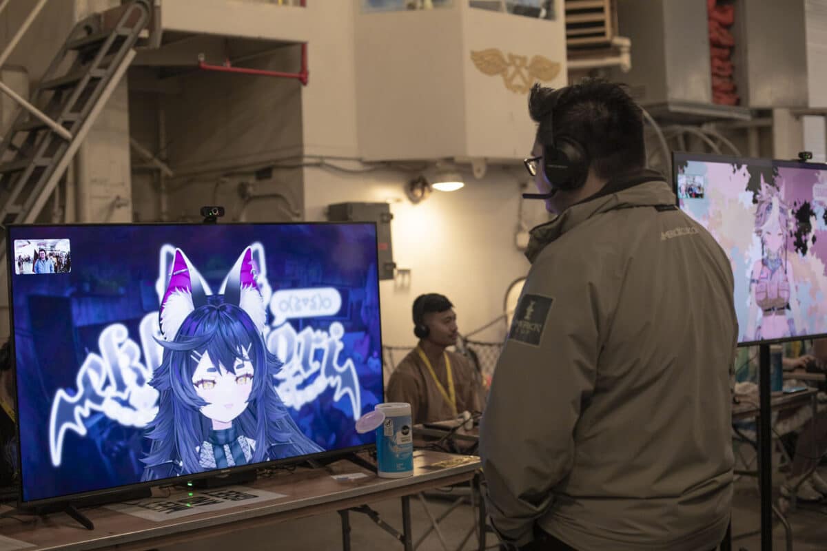Alameda Post - A man wears a headset and stands facing a screen displaying an avatar of a vtuber. they are talking.