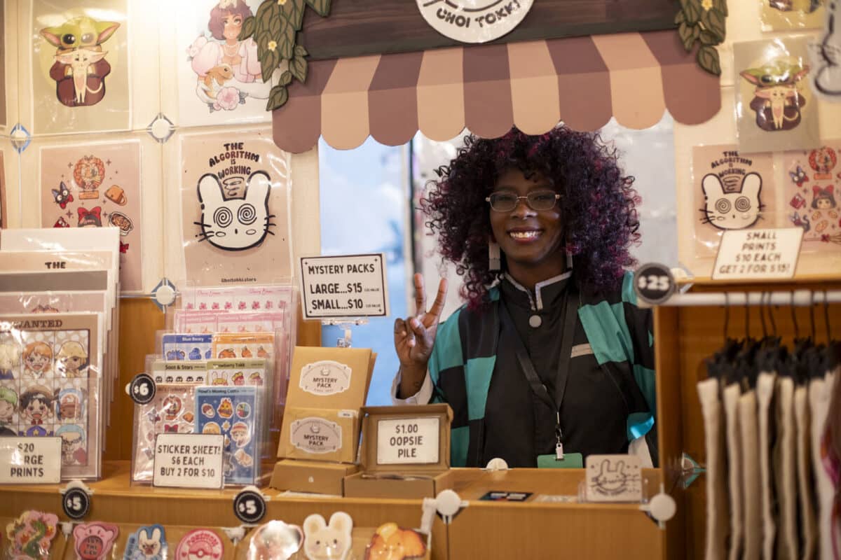 Alameda Post - A woman with curly hair stands in a very cutely decorated vendor booth and smiles and flashes a peace sign.