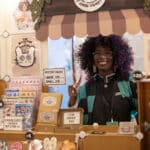Alameda Post - A woman with curly hair stands in a very cutely decorated vendor booth and smiles and flashes a peace sign.