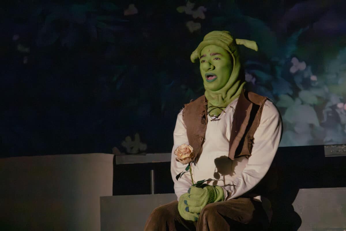 Alameda Post - The Shrek actor in full costume and makeup sits onstage, holding a rose, and sings with a serious expression.