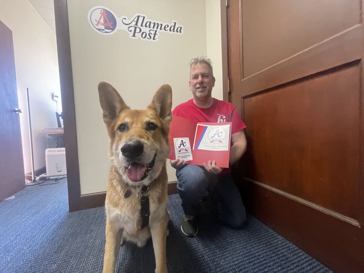 Alameda Post - Mouf the dog sits in the Alameda Post office. Adam Gillitt sits behind him holding an Alameda Stars certificate. 