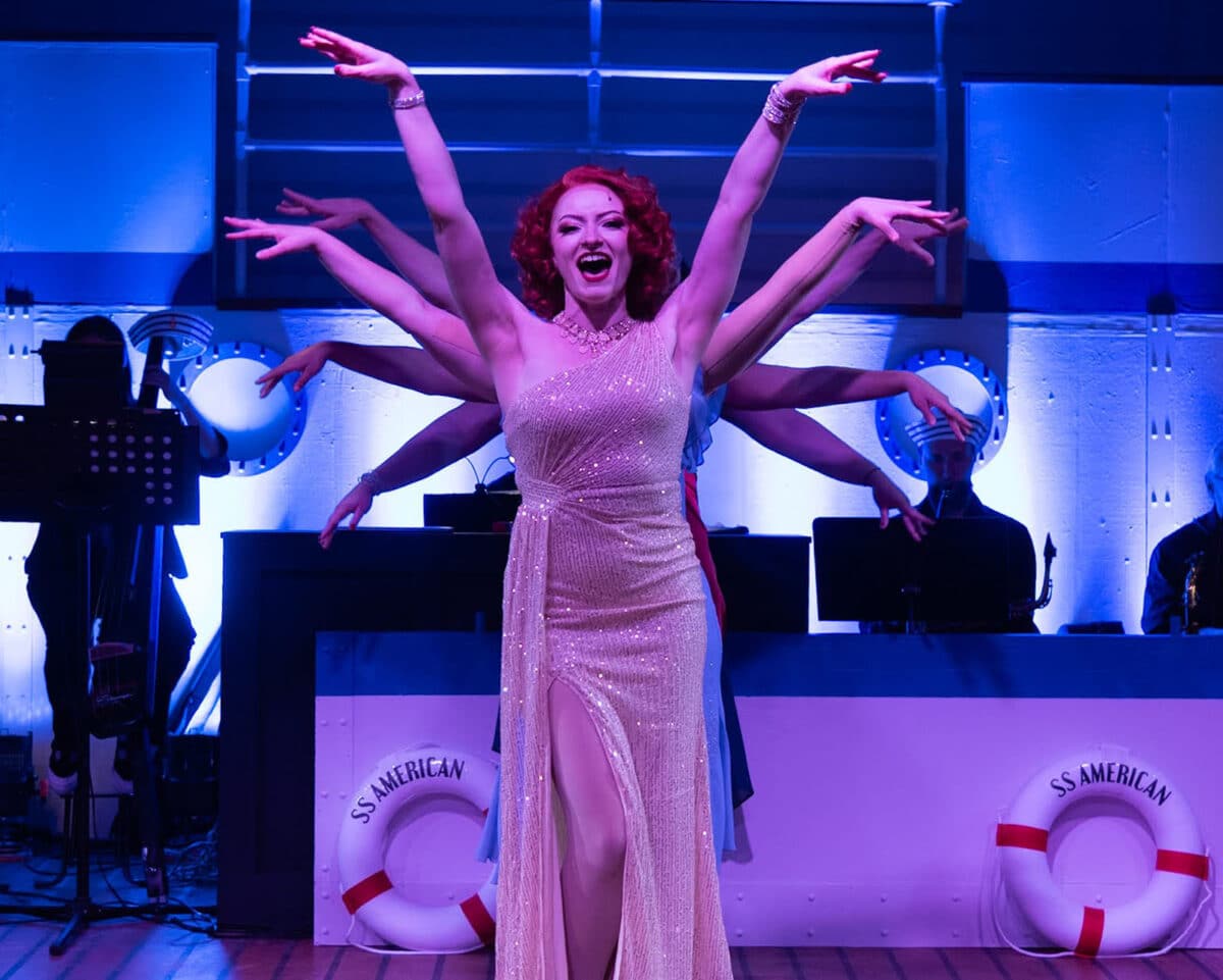 Alameda Post - Actors dance onstage in Anything Goes. They are all posed together so only a woman in front is visible, and all the people behind her stretch out their arms out to the sides in a fan behind her.