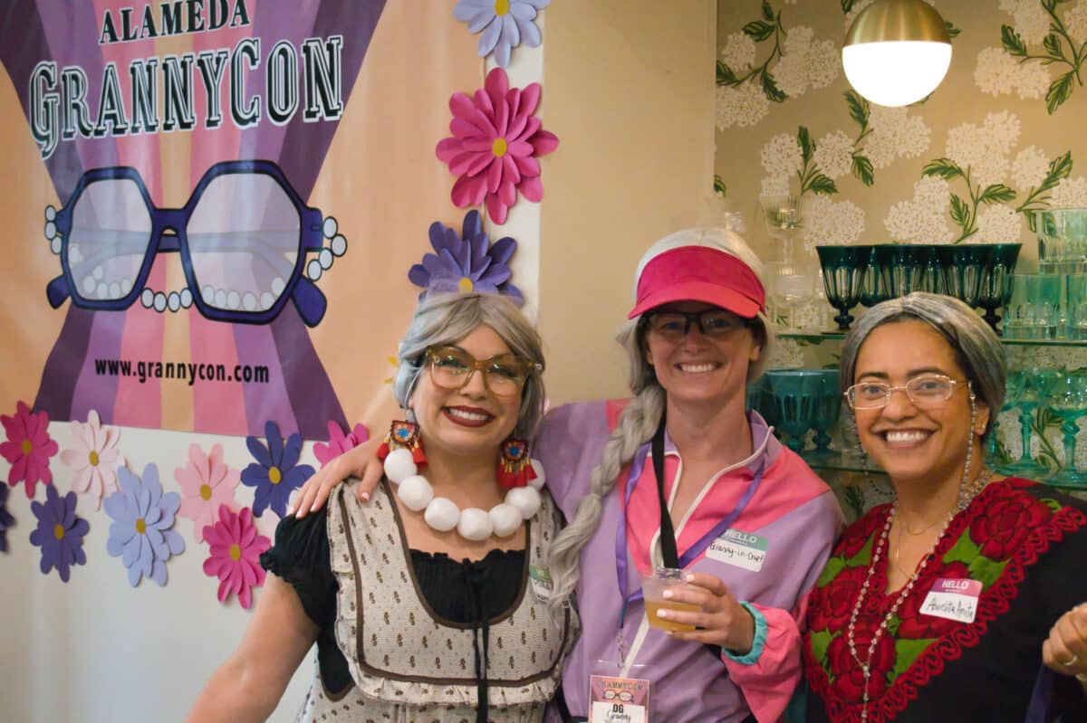 Alameda Post - Three women dressed as grannies smile next to an Alameda GrannyCon sign. 