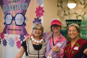 Alameda Post - Three women dressed as grannies smile next to an Alameda GrannyCon sign.