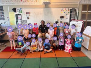 Alameda Post - A smiling adult sits with a class full of elementary schoolers with flower emojis over their faces.