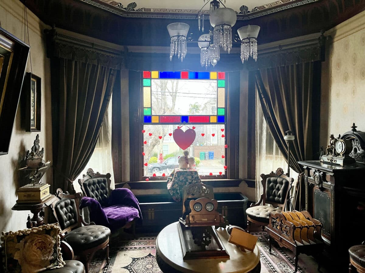 Alameda Post- A parlor in the 1890s Victorian-era style, with rich furnishings, heavy picture frames, and a chandelier.