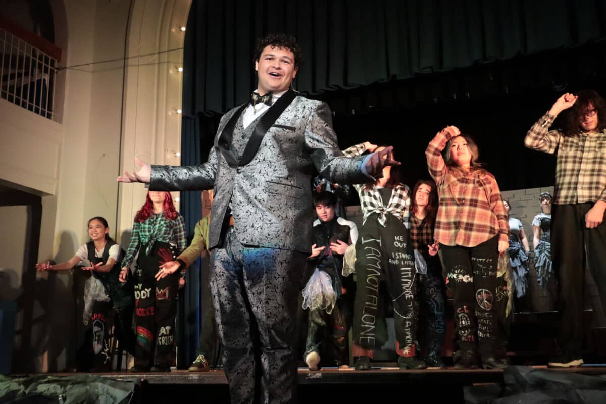 Alameda Post - A performer in a shiny silver and black suite performs in front of background of ensemble characters in Hadestown Teen Edition at Saint Joseph Notre Dame High School. 