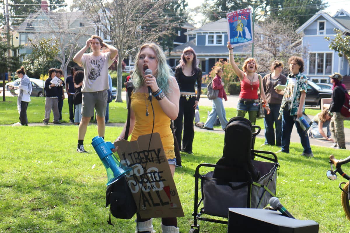 Alameda Post - A student speaks into a microphone while others behind them wave signs and make exclamations. 