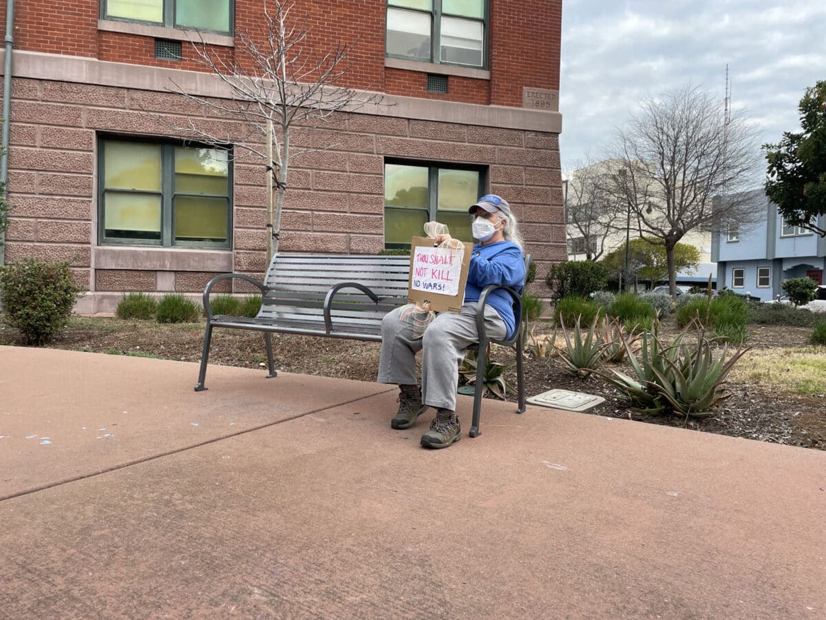 Alameda Post - A woman sits on a bench at City Hall. She wears a mask and holds a sign that says "Thou shalt not kill. No wars!"