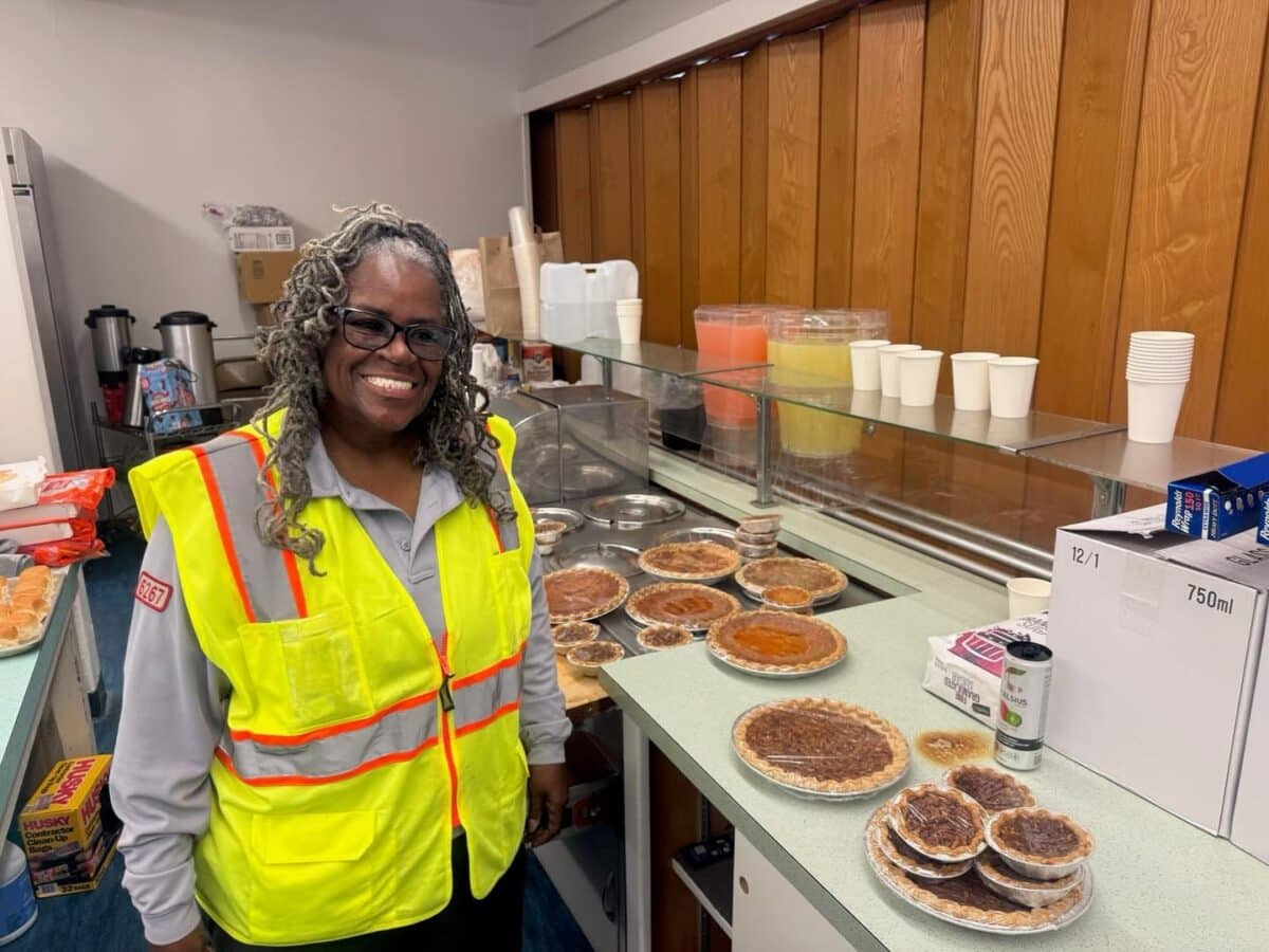 Alameda Post - A woman in a high visibility vest stands and smiles next to a counter full of pies. 