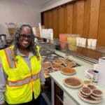 Alameda Post - A woman in a high visibility vest stands and smiles next to a counter full of pies.