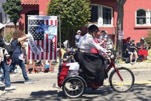 Alameda Post - A woman rides a tricycle in the Fourth of July parade. She had a No Kings sign on the back.