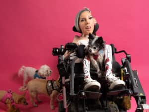 Alameda Post - A woman poses in front of a bright pink background with five dogs. She wears brightly colored accessories and has short silver hair and tattoos. She sits in a power wheelchair, and a dog sits in her lap.