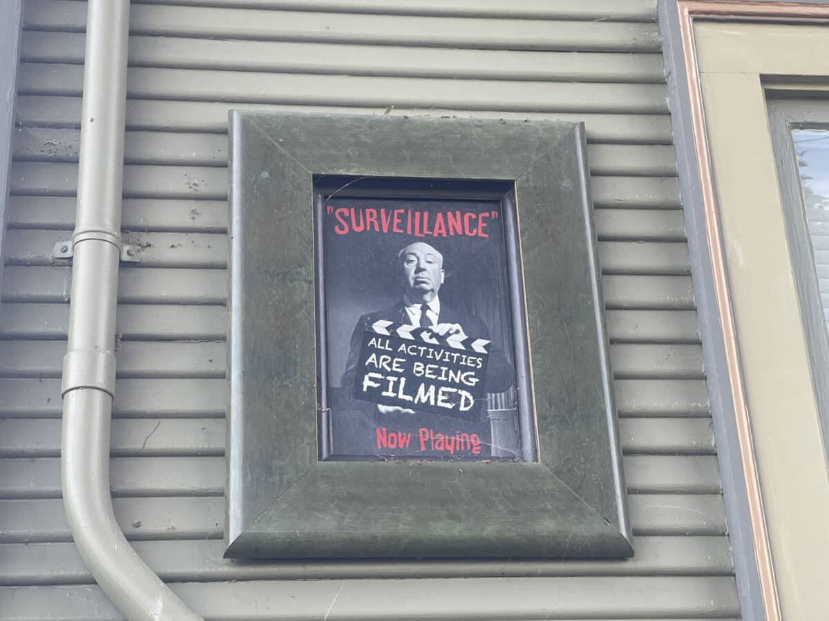 Alameda Post - An artwork hung on the outside of a home. In the frame is a black and white photo of a man holding a clapperboard. Text reads, "Surveillance" all activities are being filmed. Now playing. 