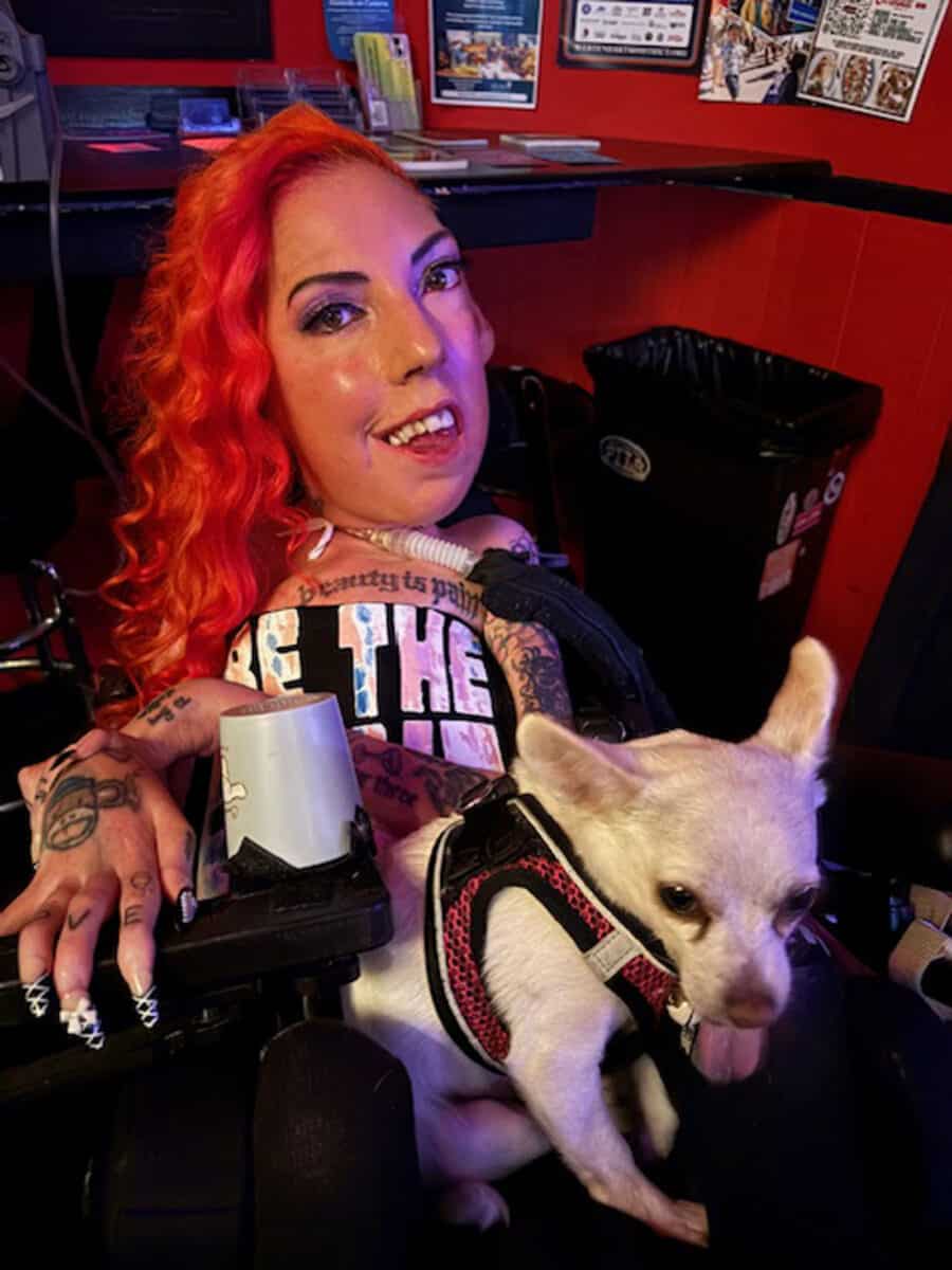 Alameda Post - Annie Wilson smiles at the camera with a small white dog on her lap. They are indoors. Annie has bright red curled hair and intricately decorated nails. A tattoo across her chest says "Beauty is pain." She is using a power wheelchair and tracheostomy tube.