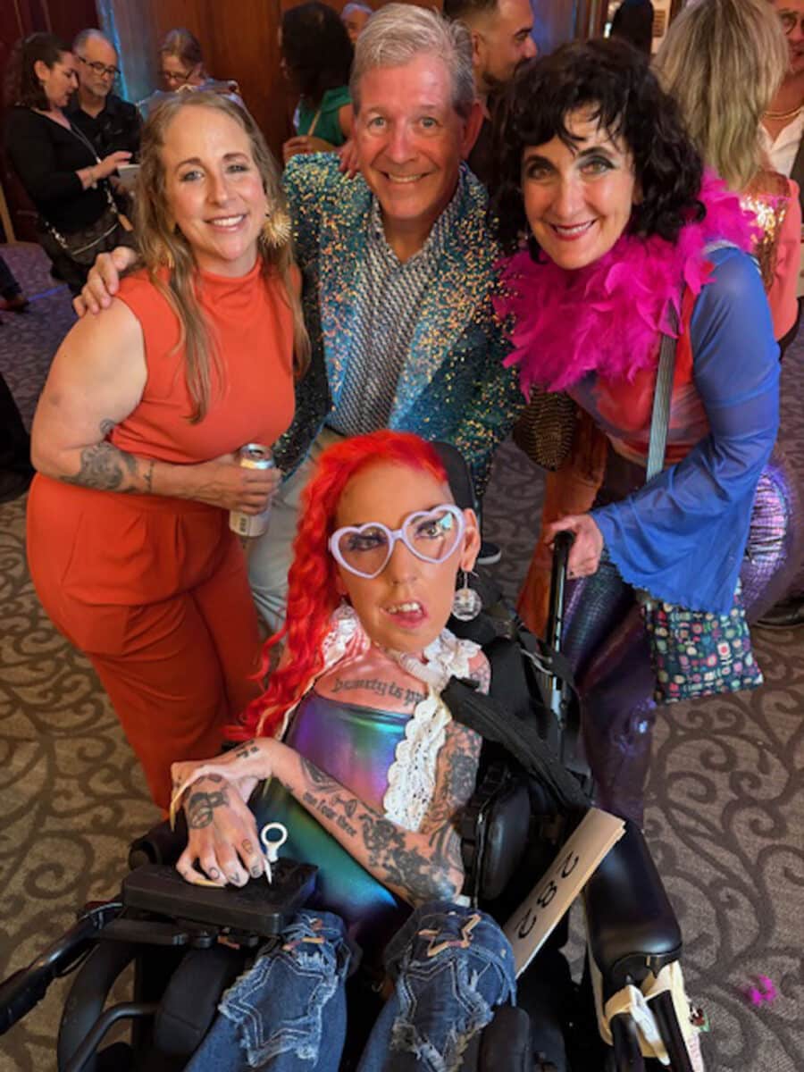 Alameda Post - Annie Wilson poses with three other people leaning in close behind her power wheelchair. She wears lavender colored heart shaped glasses, bold clothing, and flaming red hair. Everyone is smiling and colorfully dressed.