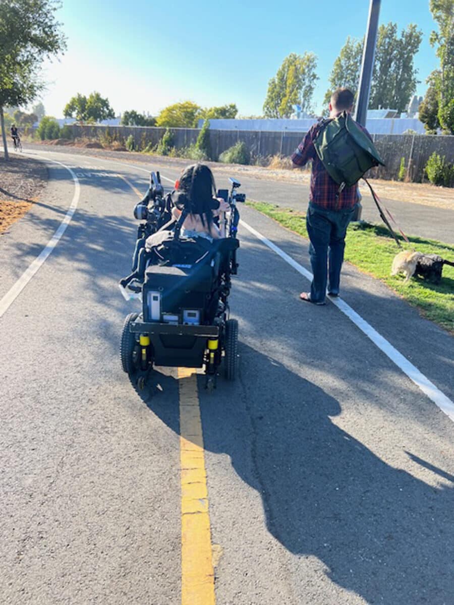 Alameda Post - Annie rolls away from the camera on a paved path at Jean Sweeney Open Space Park. Next to her, a man carries a bag and walks two dogs.