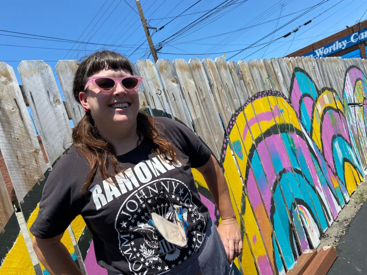 Alameda Post - a woman in sunglasses stands in front of a mural painted on a fence