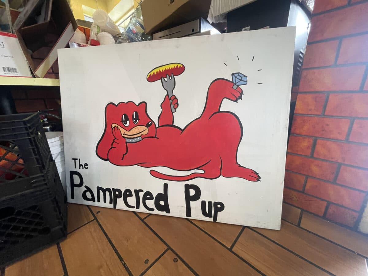 Alameda Post - A hand painted sign inside of a restaurant that says "The Pampered Pup and has an image of a mascot dog holding a hotdog. 