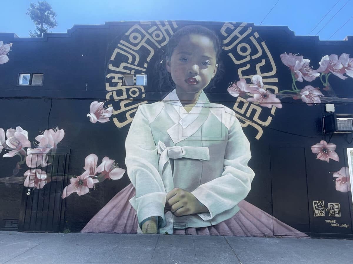 Alameda Post - A mural of a young mixed race girl wearing a hanbok, a traditional Korean dress, surrounded by cherry blossoms. 