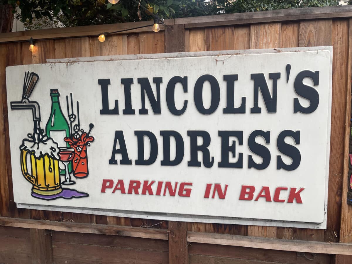 Alameda Post - A large sign on the fence that has images of adult beverages and words that say "Lincoln's Address. Parking in back."