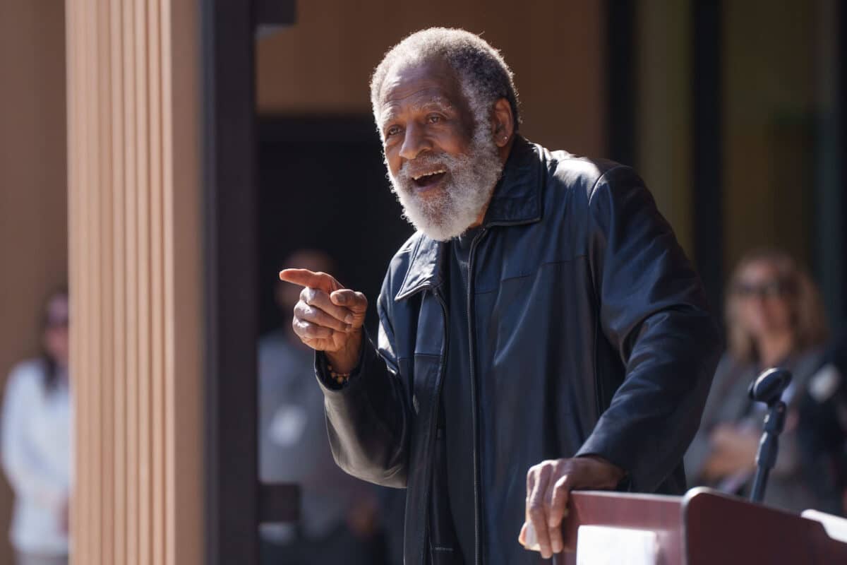Alameda Post - A gentleman with greying hair and a beard smiles and points while leaning on a podium. The man is Arnold Perkins.