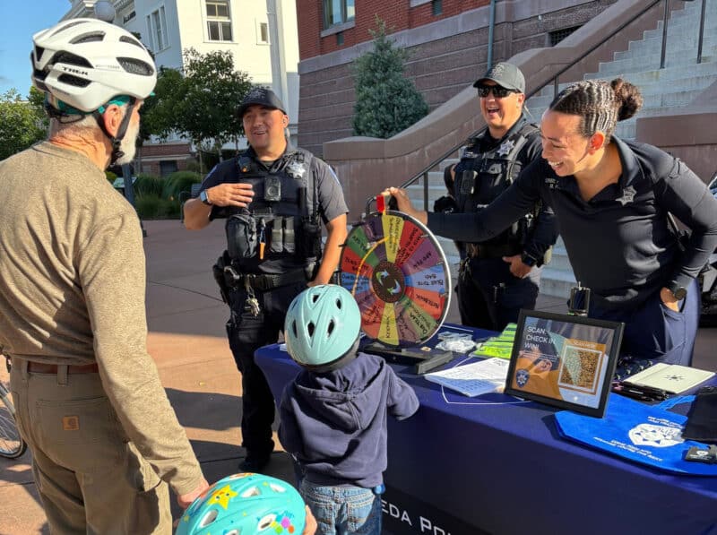 Alameda Post - A.P.D. officers outside of City Hall stand at a booth and talk to community members wearing bike helmets.