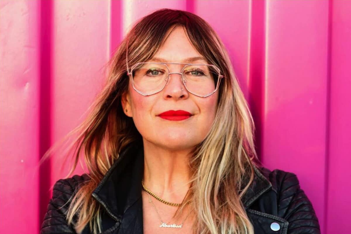 Alameda Post - A headshot of Kate Schatz wearing a leather jacket, bright lipstick, and a necklace that says abortion. 