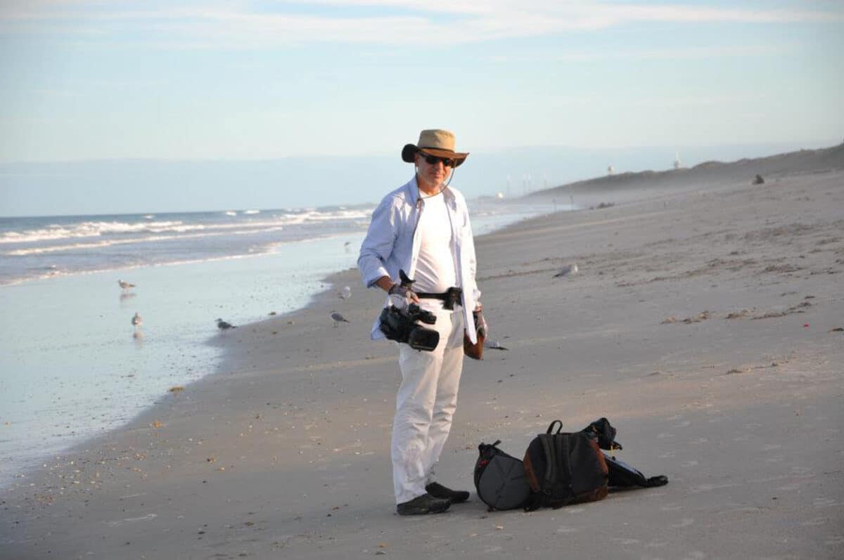 Alameda Post - A man stands on the beach in a hat, holding a camera.