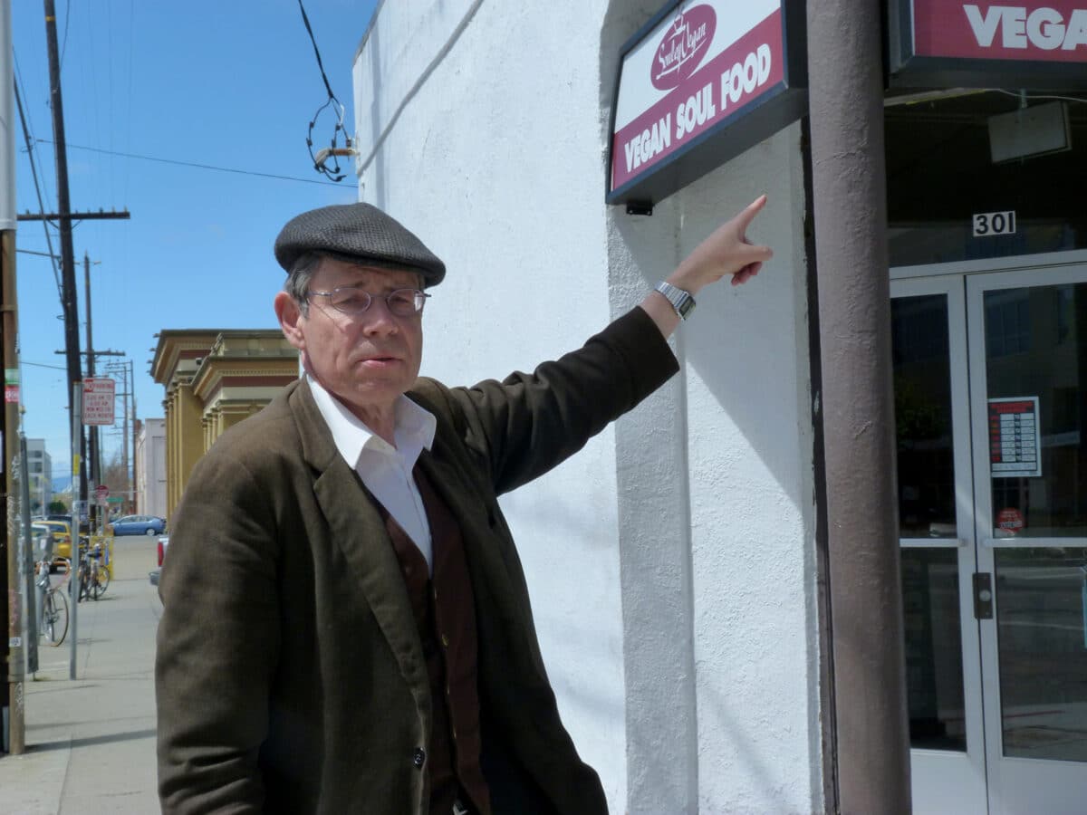 Alameda Post - Christopher Buckley points to a building with a Vegan Soul Food sign in front.