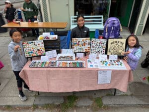 Alameda Post - Three sisters stand surrounding a table full of small crafts including braided keychains and cute ceramic animal or fruit shaped worry stones.
