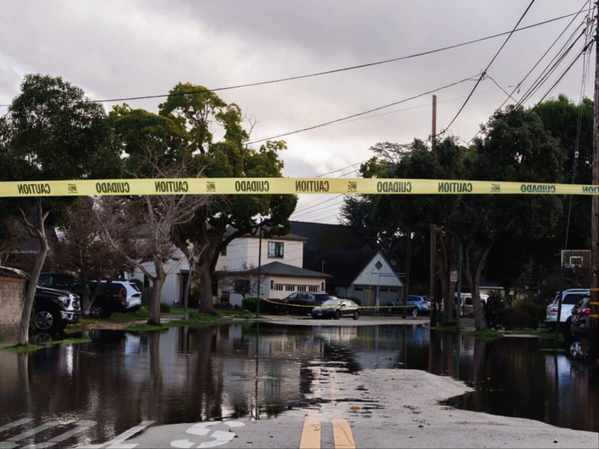 Alameda Post - A piece of caution tape across an intersection. Flooding on an Alameda street is visible in the background