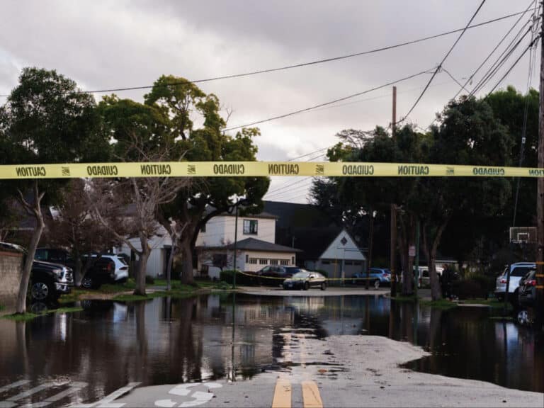 Alameda Post - A piece of caution tape across an intersection. Flooding on an Alameda street is visible in the background