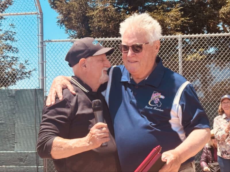 Alameda Post - Little League President Ron Matthews hugs another man in front of a crowd at the ball field.