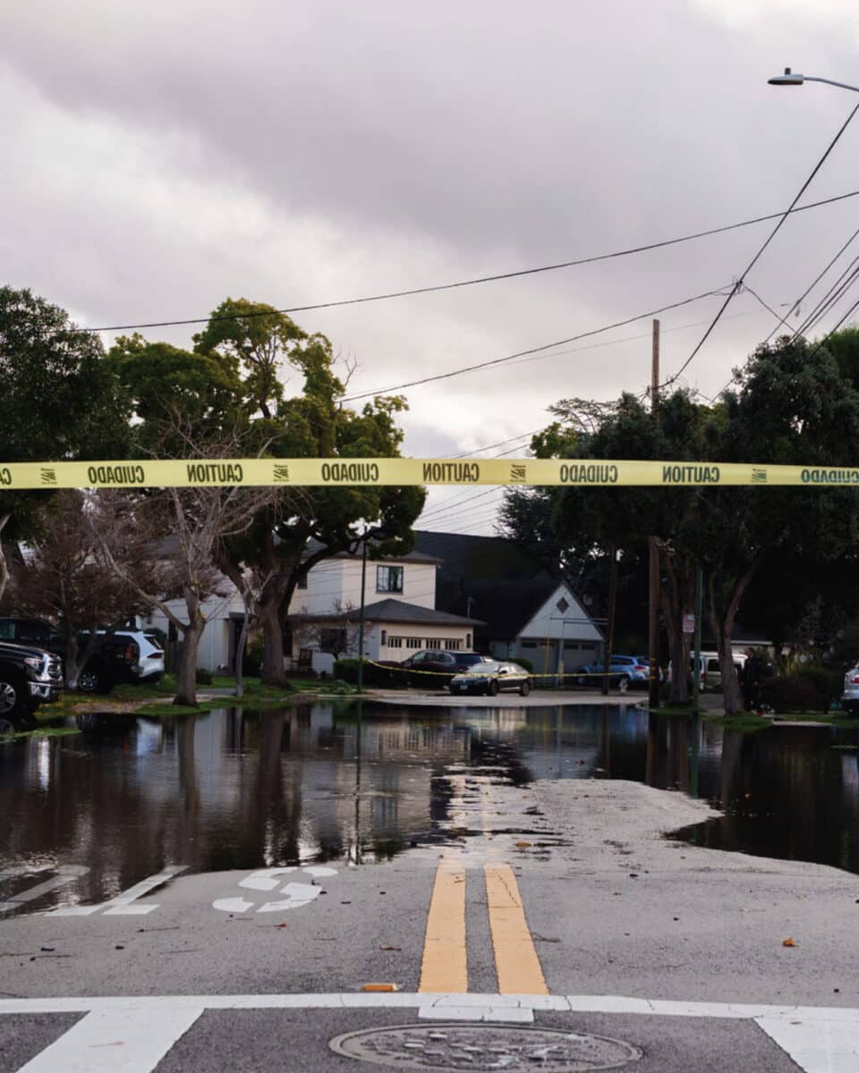 Alameda Post - A piece of caution tape across an intersection. Flooding on an Alameda street is visible in the background 