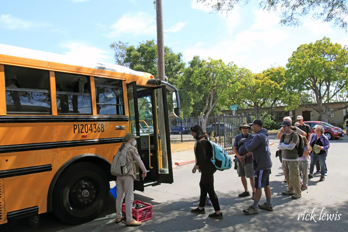 Alameda Post - A group of people line up to board a school bus. 