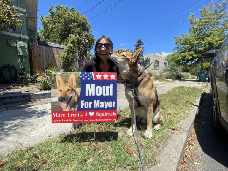 Alameda Post - Mouf the dog gives a kiss to a woman. They sit or crouch together next to a Mouf for Mayor yard sign by the road.