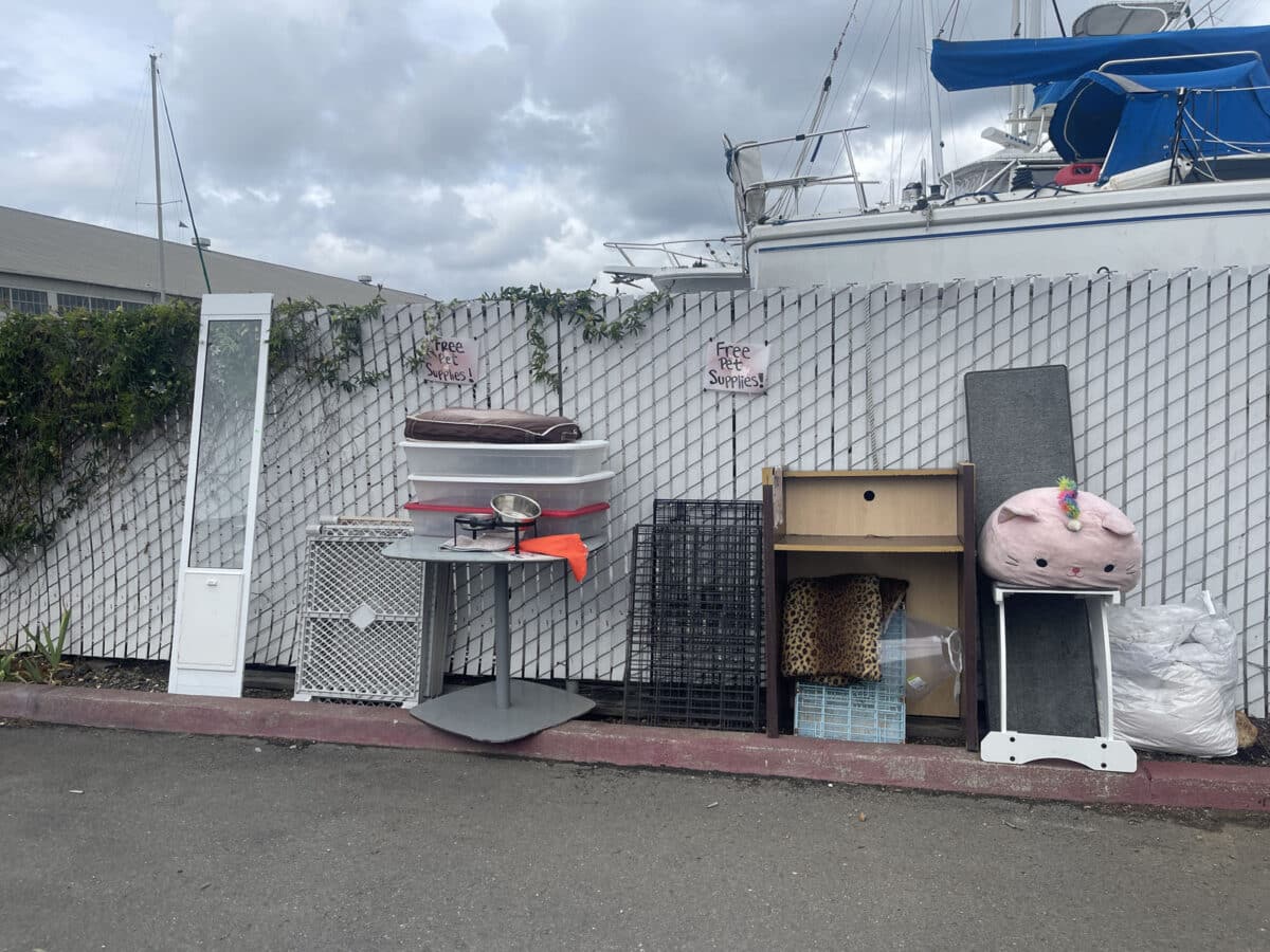 Alameda Post - A somewhat organized assortment of items lined up outside by a fence. The items include furniture, dog crates, accessories, and bedding. 