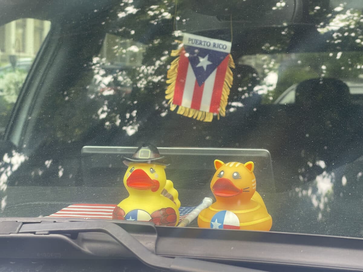 Alameda Post - A photo of the a car dashboard taken from outside the car. A small Puerto Rico flag hangs from the rearview mirror, and two Puerto Rico rubber ducks sit under it. 
