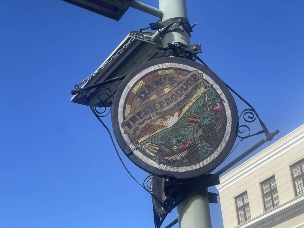 Alameda Post - A circular colorful sign that appears to be made of wood. The sign has fruits and vegetables and farmland on it, and it says "Dan's Fresh Produce. Like Having a farmers market everyday."