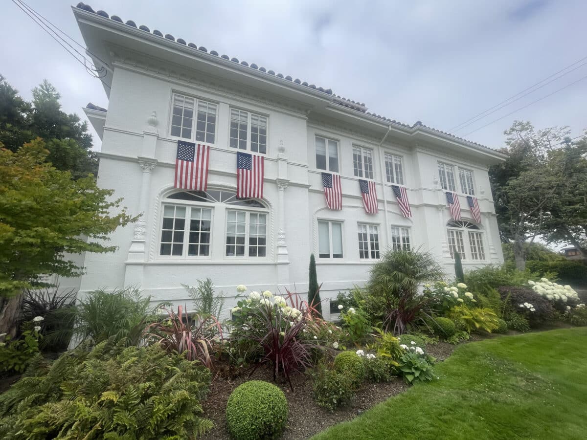 Alameda Post - The side of a grand white house with seven American flags draped around the side. 
