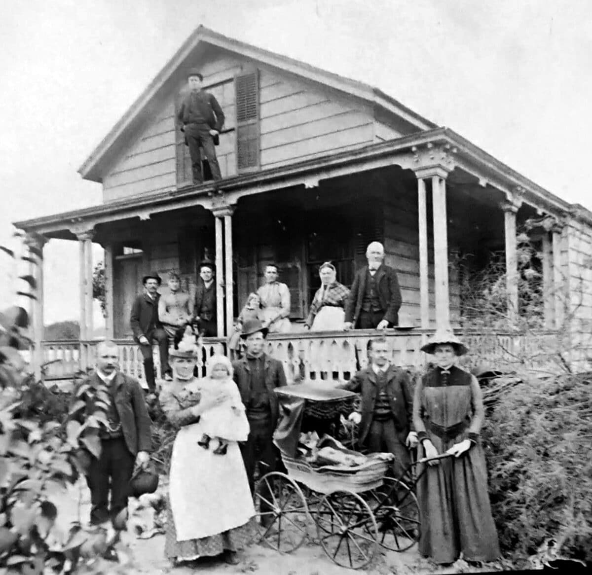 Alameda Post - A black and white family photo of twelve adults, two children, and a baby stroller in front of a two story wooden house with a porch. 