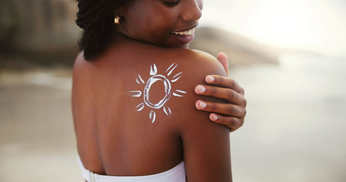 Alameda Post - A woman with dark skin standing at the shoreline smiles over her shoulder and looks at sunscreen shaped like a sun on her right shoulder. 