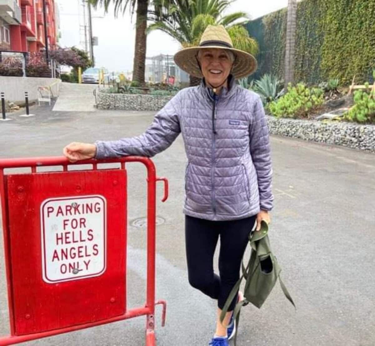 Alameda Post - Holly Larsen stands next to a gate in a parking lot that says Parking for Hells Angels only. She is wearing comfortable clothes and a hat, and she leans on the gate and smiles. 