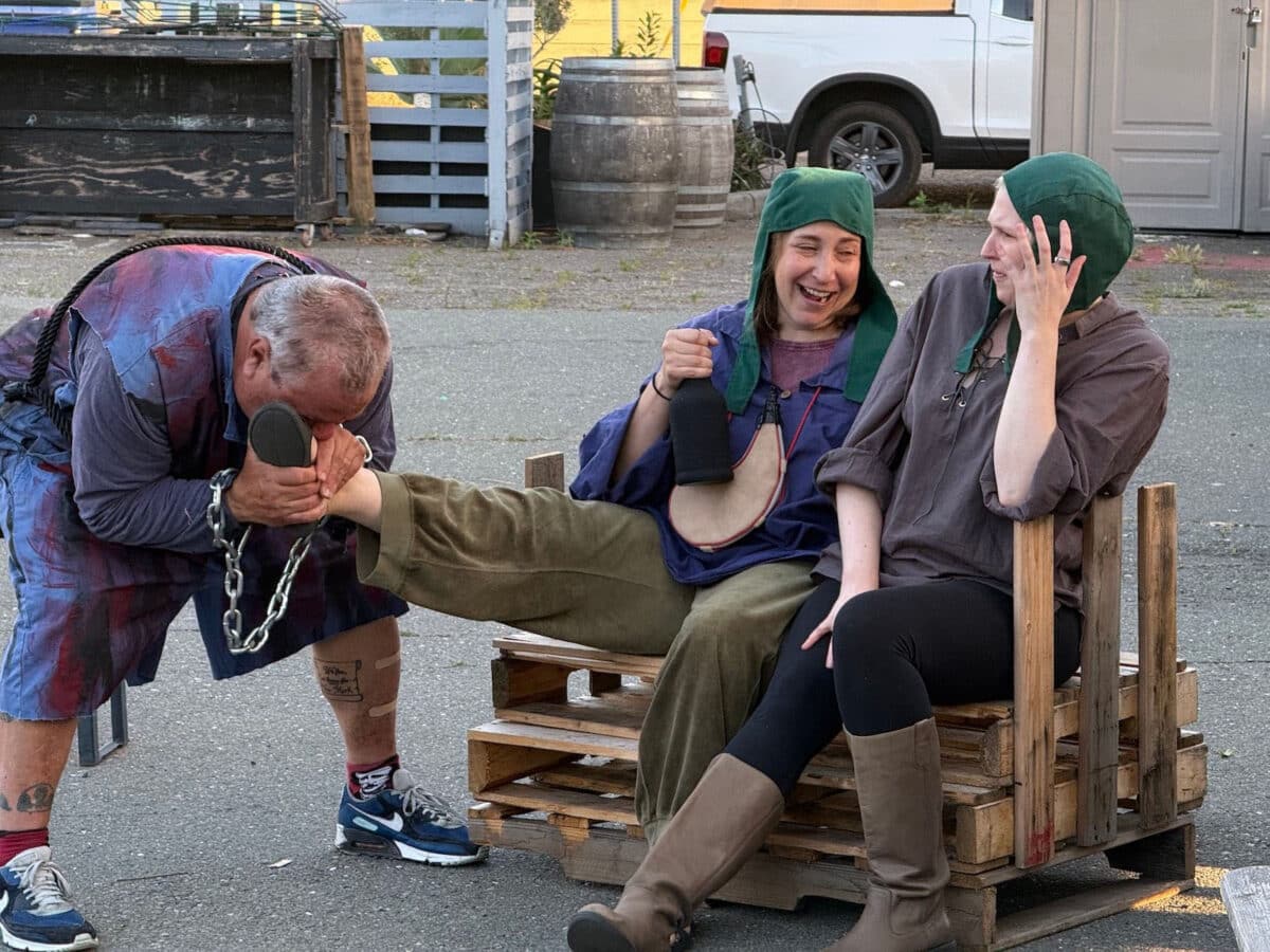 Alameda Post - Two actors sit together and laugh. One holds a flask and they appear to be playing comic characters.Another actor holds one of their ankles and appears to kiss it.
