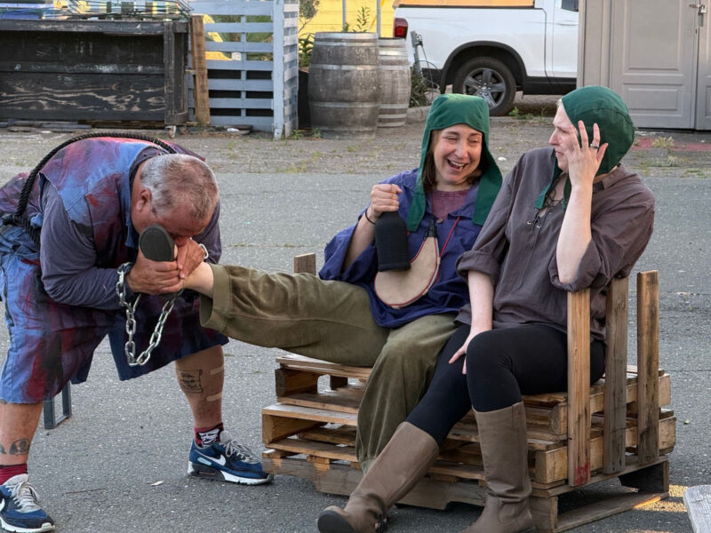Alameda Post - Two actors sit together and laugh. One holds a flask and they appear to be playing comic characters.Another actor holds one of their ankles and appears to kiss it.